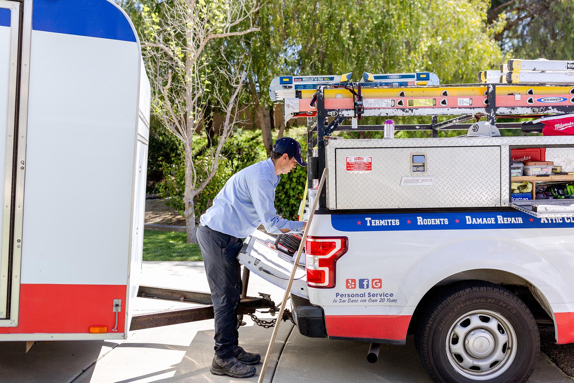 Pest control technician preparing equipment from a service truck labeled with tools and ladders, parked in a residential driveway.