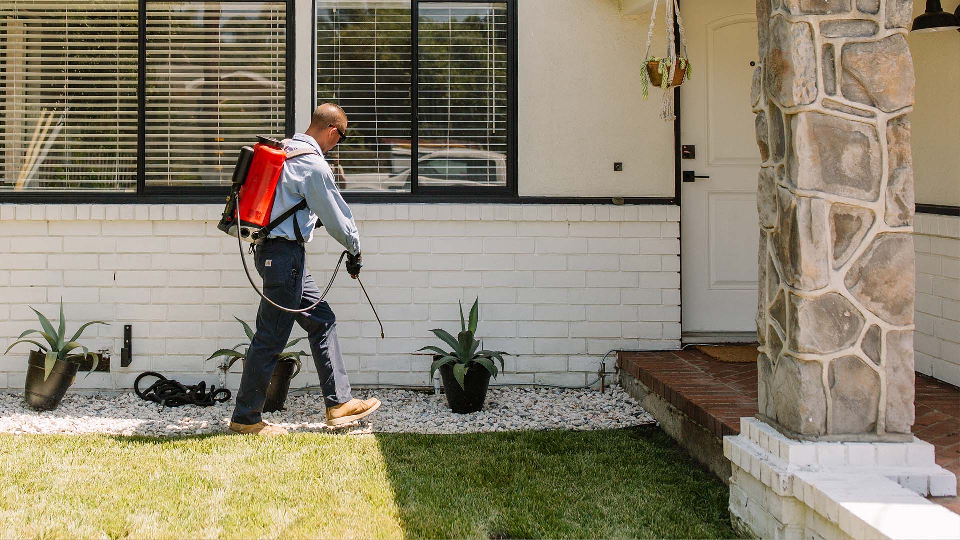 Pest control technician wearing a red backpack sprayer and treating the exterior of a house near the front entrance.