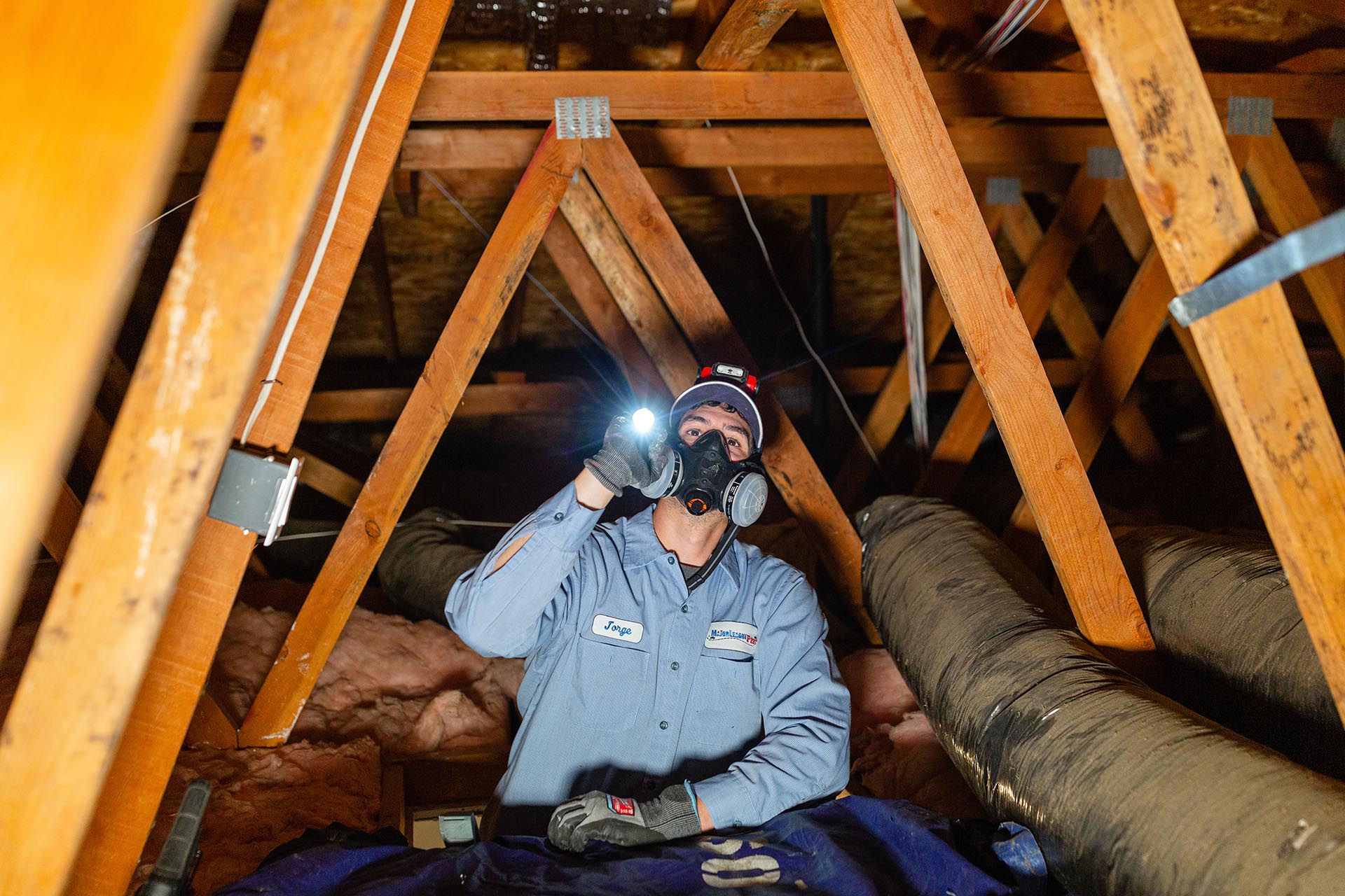 Pest control technician inspecting a residential attic with a flashlight while wearing a protective mask and uniform.